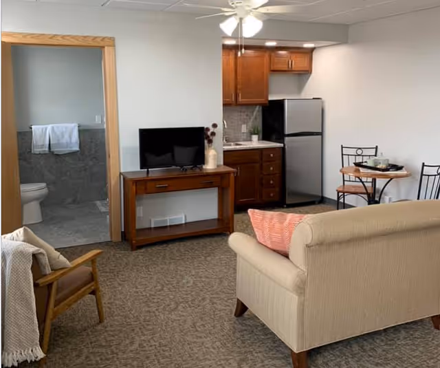 Small living area with a sofa, TV on a console, kitchenette with a refrigerator, a small dining table, and an open doorway to a bathroom.