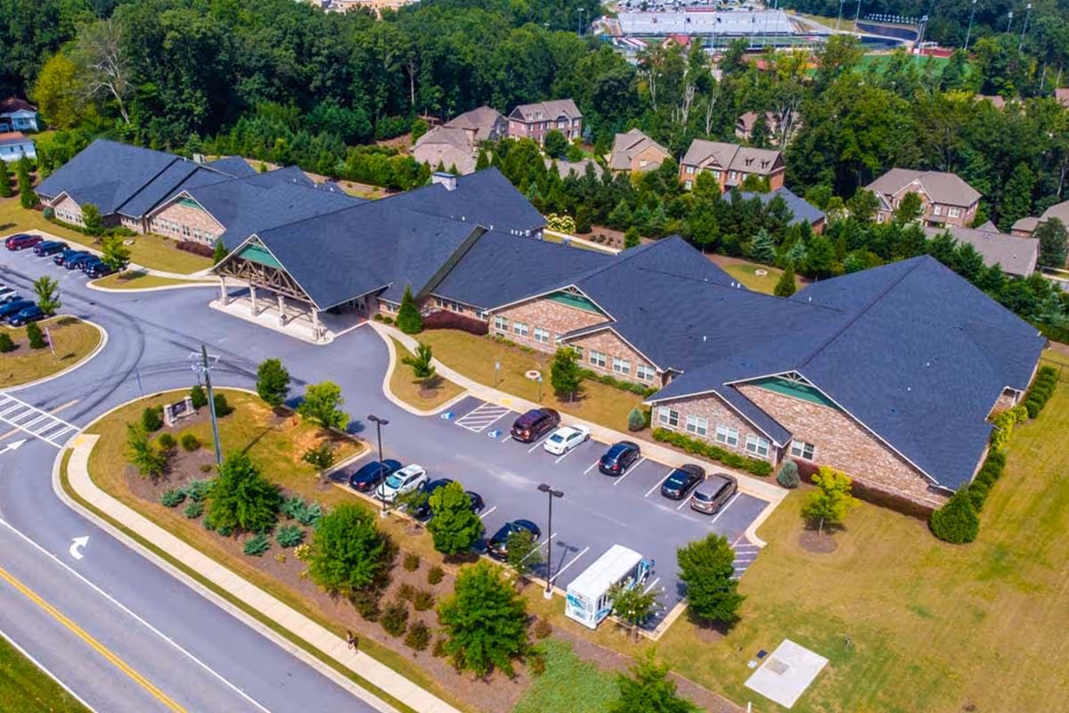 Aerial view of Antebellum James Burgess senior living facility showing a large single-story brick building with a dark roof, surrounded by landscaped greenery, parking spaces with several cars, and a driveway leading to the main entrance with a covered drop-off area. Residential houses and trees are visible in the background.