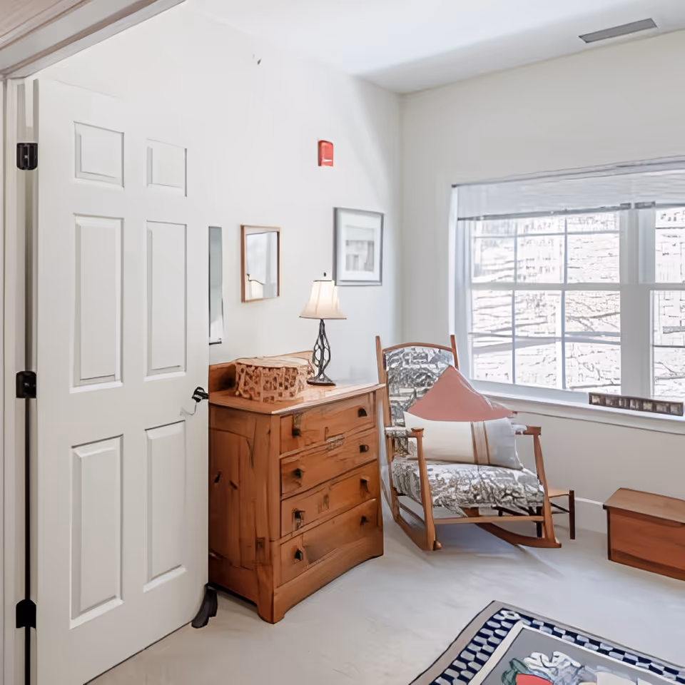 Bright sitting area with a wooden dresser, lamp and a rocking chair by a large window.