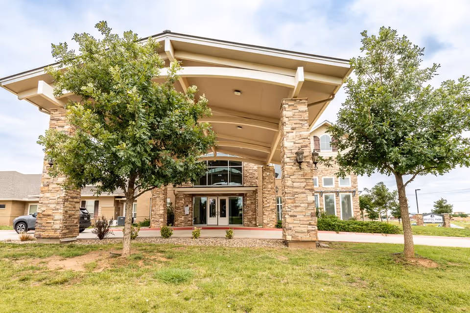 Front exterior view of Bristol Park at Amarillo Assisted & Living Memory Care building with a covered entrance supported by stone pillars, two trees in the foreground, and a grassy area.