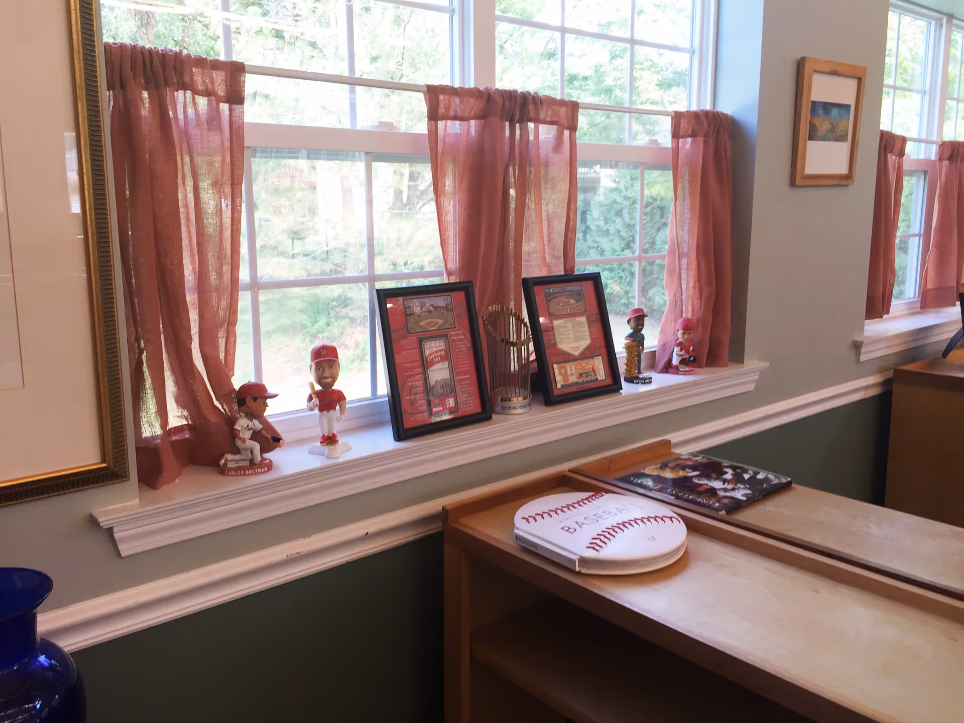 A room with two large windows adorned with sheer reddish curtains. On the windowsill are four baseball player bobblehead figurines, two framed pictures, and a small replica of a baseball trophy. Below the windowsill is a wooden cabinet with a baseball-themed book and another book on top. The walls are painted light blue above white trim and green below.