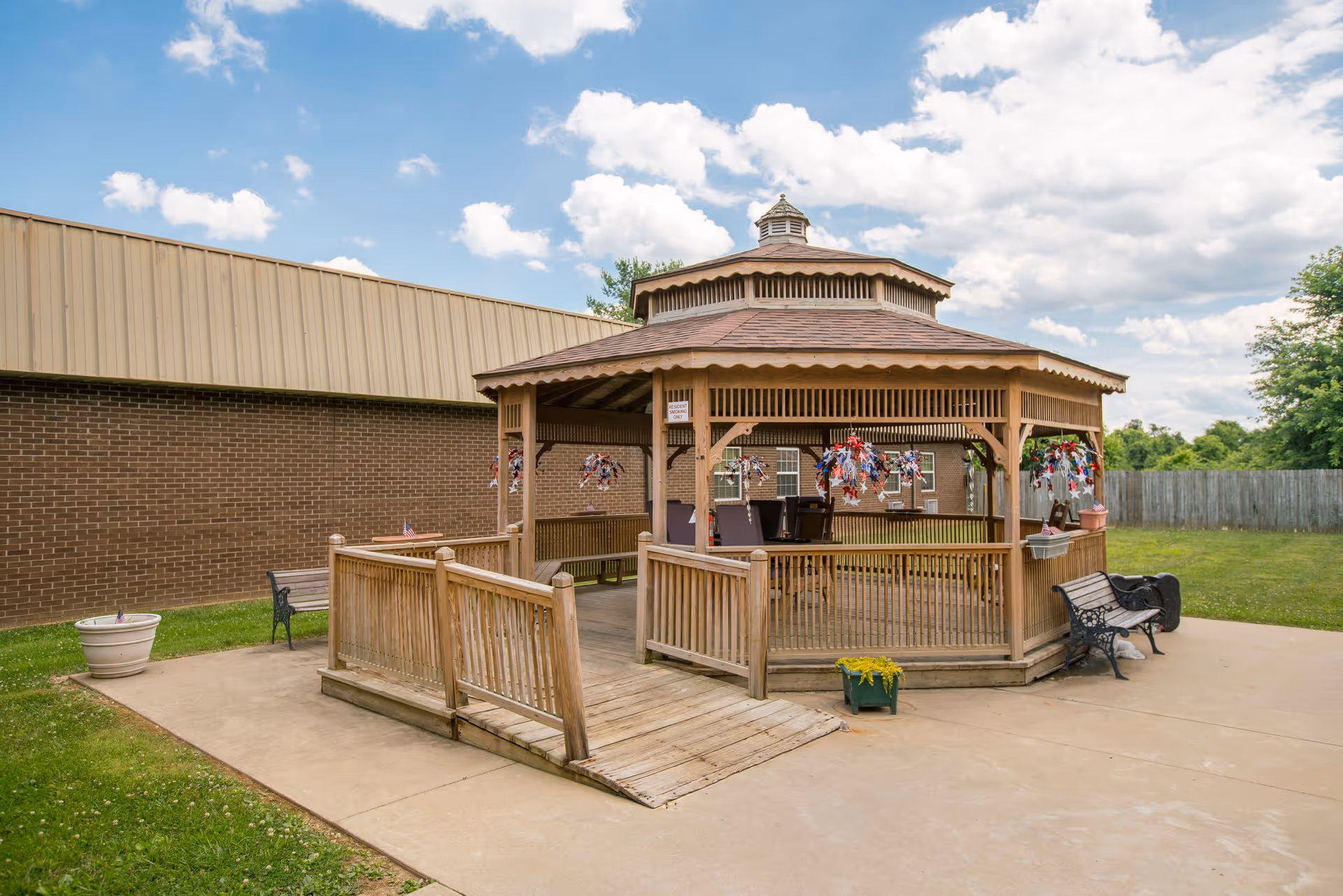 Outdoor wooden gazebo with a ramp and benches around it, decorated with red, white, and blue hanging ornaments. The gazebo is situated on a concrete patio next to a brick building, with a grassy area and a wooden fence in the background under a partly cloudy sky.