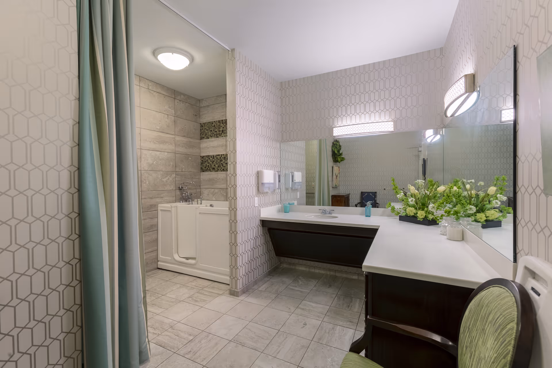 A spacious bathroom featuring a walk-in bathtub with tiled walls and a curtain for privacy. The room has a large countertop with a sink, a mirror spanning the wall above the counter, and decorative flowers placed on the counter. The floor is tiled, and there is a green cushioned chair near the counter.