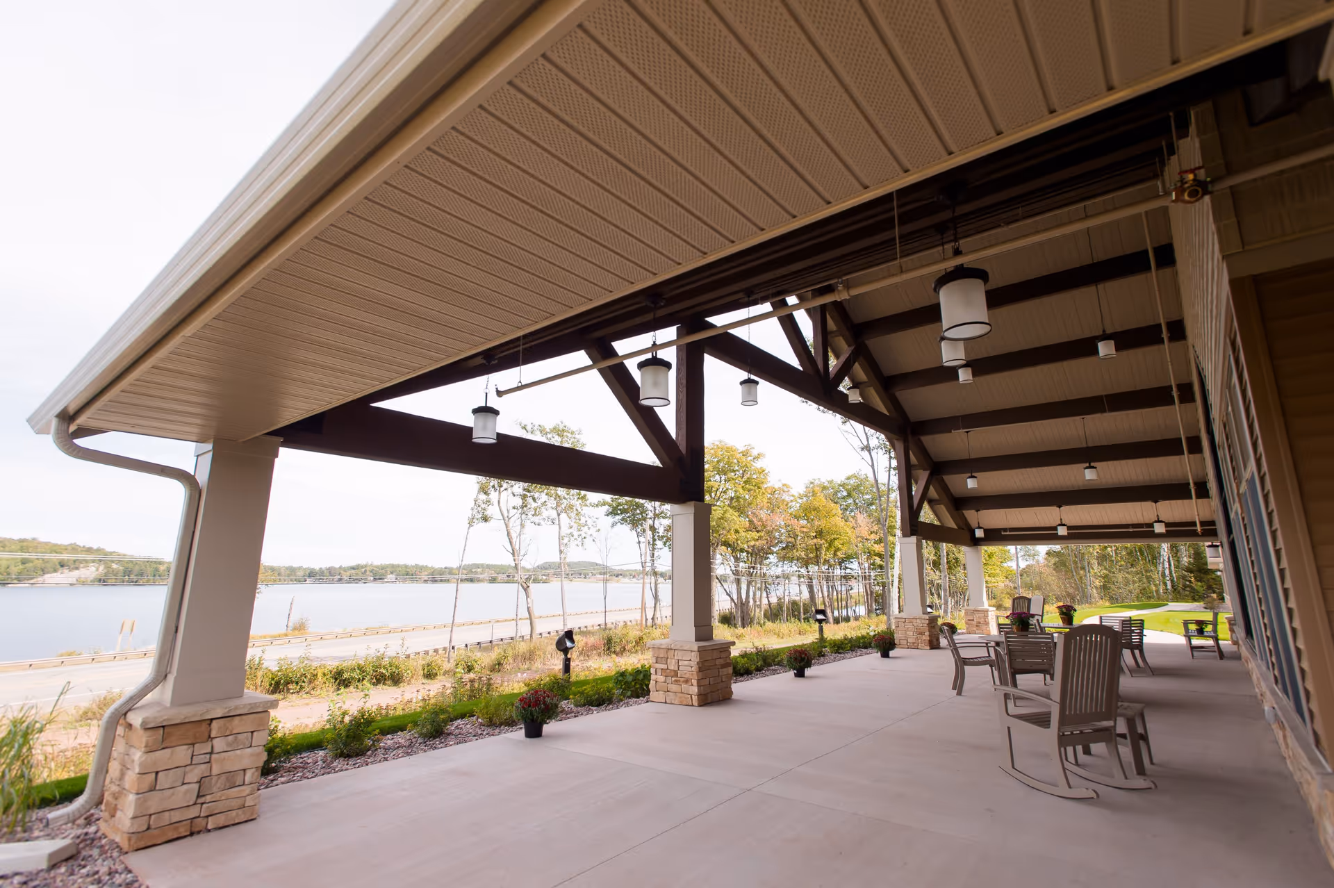Covered outdoor patio area with hanging lantern-style lights, stone pillars, and several chairs and tables. The patio overlooks a scenic view of a lake, trees, and a road.