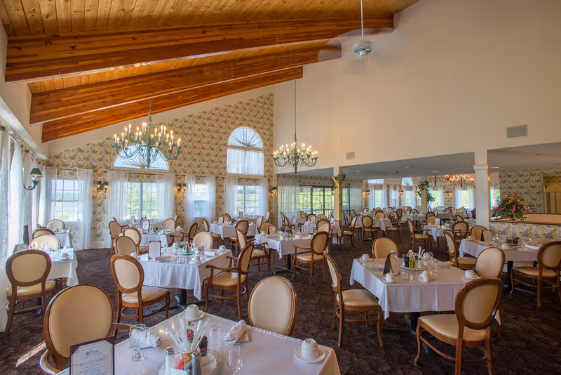 A spacious dining room with multiple tables covered in white tablecloths, each set with glassware, napkins, and condiments. The room features high vaulted wooden ceilings with exposed beams, large windows with sheer curtains allowing natural light, and elegant chandeliers hanging from the ceiling. The walls have patterned wallpaper and the floor is carpeted.