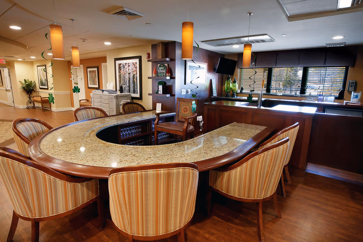 Interior view of a senior living facility's common area featuring a curved granite countertop bar with striped upholstered chairs around it. The space is warmly lit with hanging pendant lights and decorated with green shamrock streamers. In the background, there is a window with blinds, a television mounted on the wall, and some decorative items on shelves.