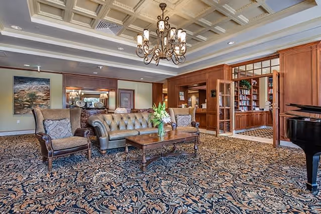 A spacious and elegant living room area with a patterned carpet, a tufted leather sofa, two upholstered armchairs, a wooden coffee table with a vase of flowers, a chandelier hanging from a coffered ceiling, and a grand piano on the right side. In the background, there is a wooden bookshelf and a framed painting on the wall.