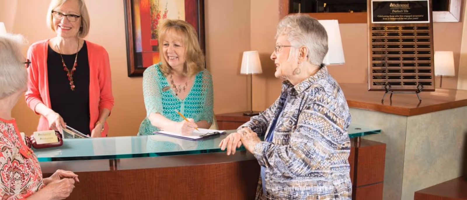 Three elderly women standing and sitting around a reception desk in a senior living facility, smiling and engaging in conversation. The reception area has a glass countertop, lamps, and a plaque on the wall behind the desk.