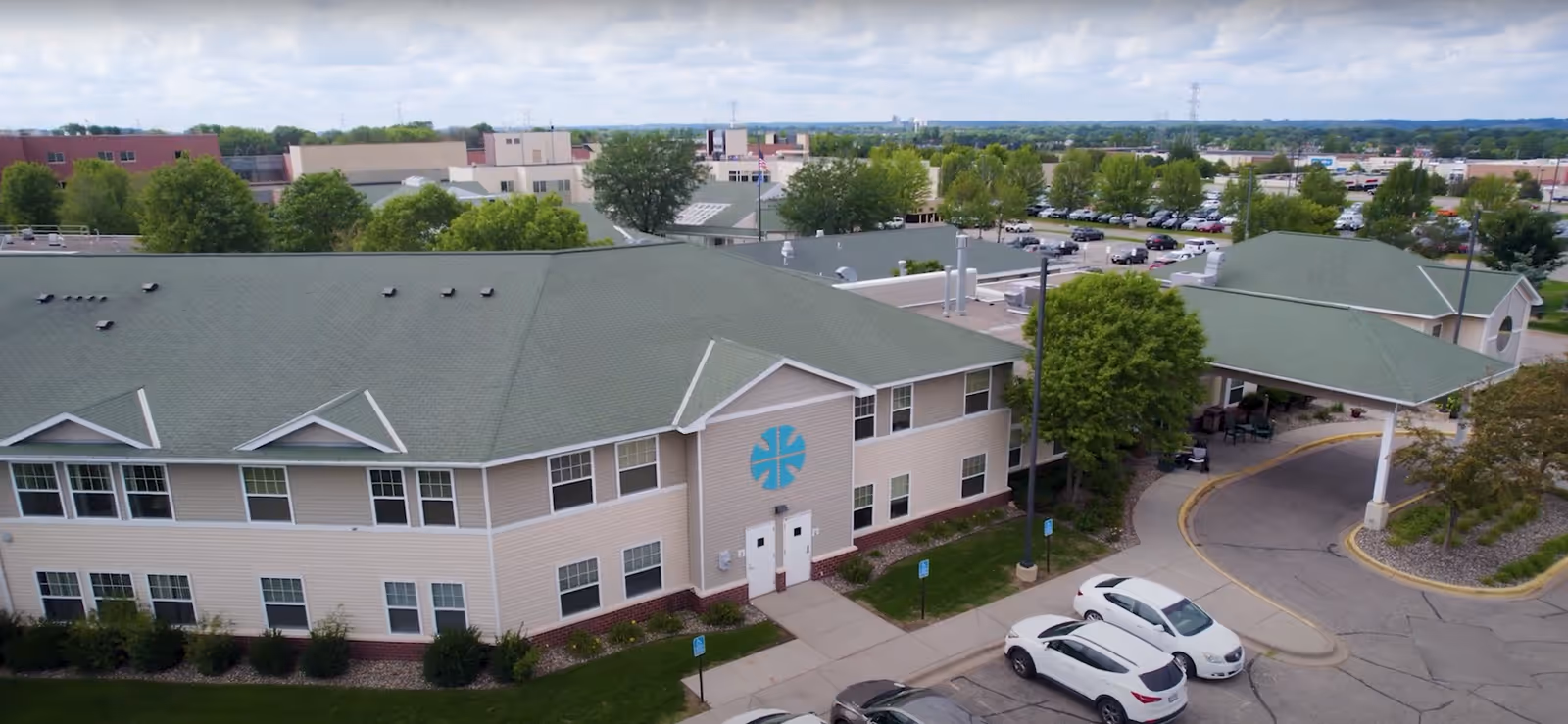 Aerial view of Benedictine Living Community-Shakopee St. Gertrude's and the Gardens building with a green roof, beige siding, and a covered entrance driveway. Several cars are parked in the parking lot, and there are trees and other buildings in the background under a partly cloudy sky.