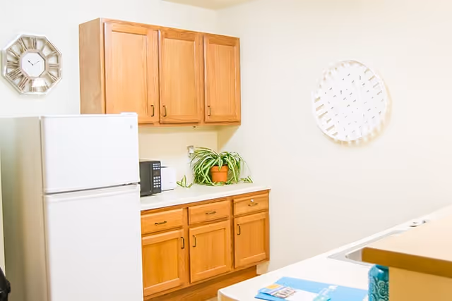 A bright kitchen area with wooden cabinets, a white refrigerator, a microwave, a potted plant on the countertop, and two decorative wall clocks on cream-colored walls.