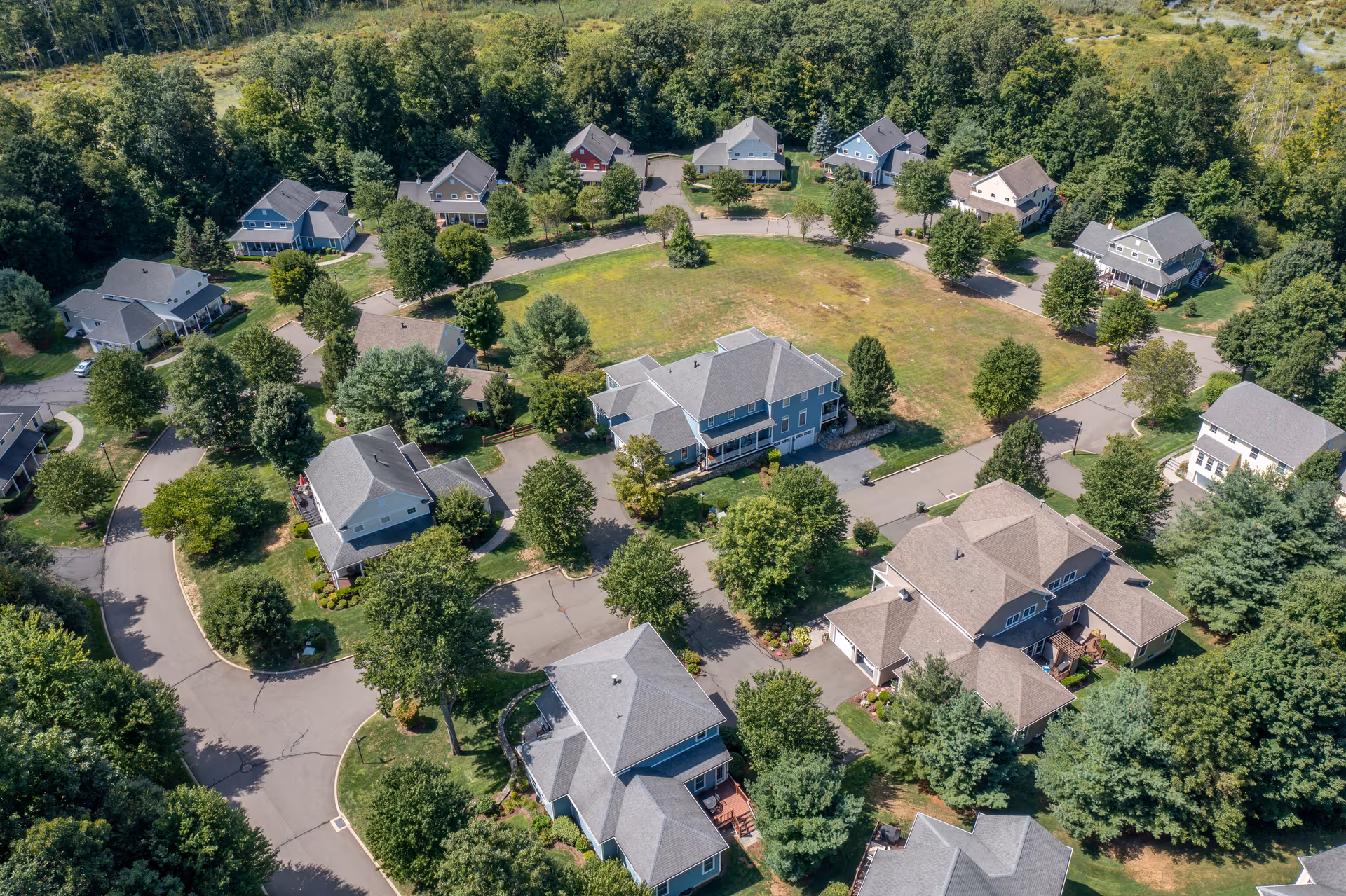 Aerial view of a residential community with multiple houses arranged around a central grassy area, surrounded by trees and greenery.