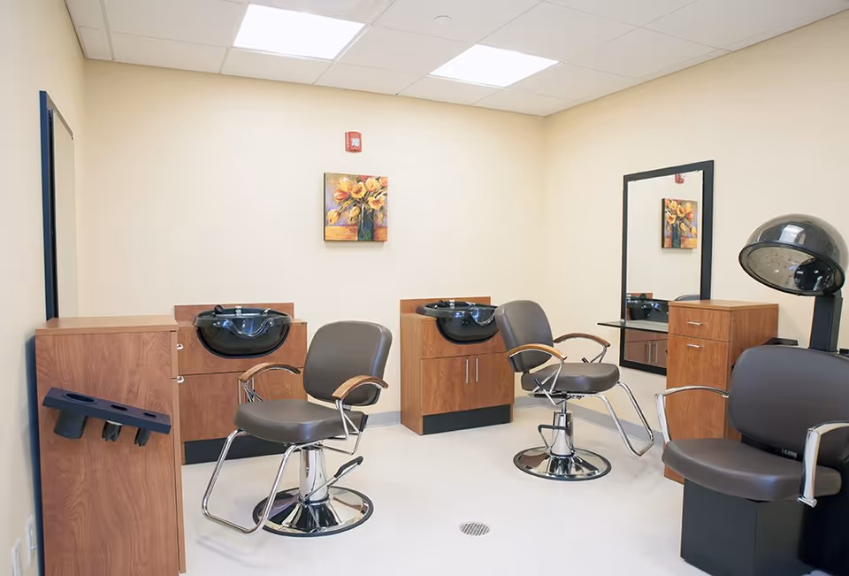 Interior of a hair salon area with two brown salon chairs in front of black wash basins mounted on wooden cabinets. There is a large mirror on the wall, a black hair dryer chair, and a small painting of yellow flowers on the wall. The room has beige walls and a white ceiling with fluorescent lights.