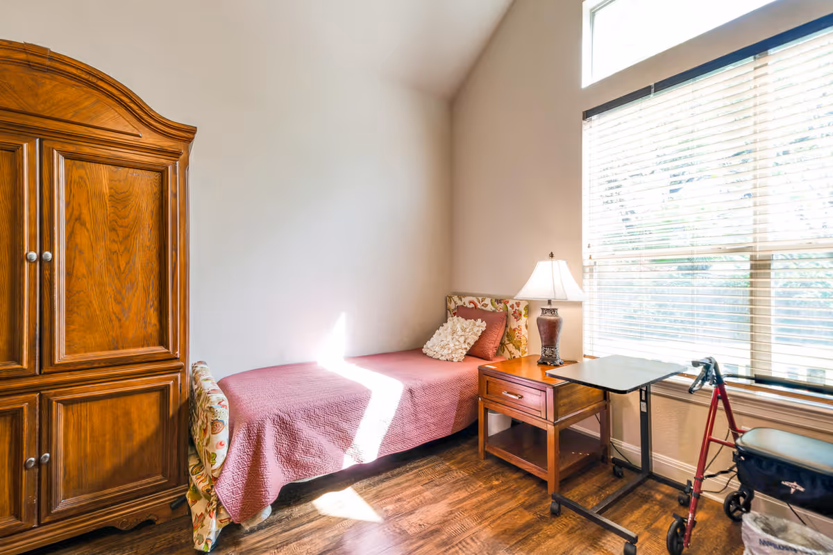 A small, cozy bedroom with a single bed covered in a pink quilt and floral headboard. Next to the bed is a wooden nightstand with a lamp and a small overbed table. A red walker is positioned near a large window with blinds, and a wooden armoire stands against the opposite wall. The room has wooden flooring and light-colored walls.