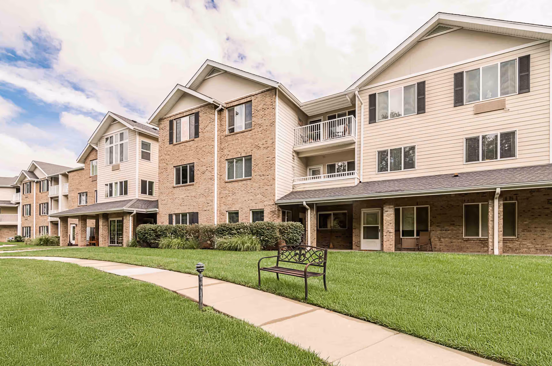 Exterior view of a multi-story senior living facility building with beige siding and brick accents. There is a well-maintained green lawn with a curved concrete walkway and a decorative metal bench. The sky is partly cloudy.