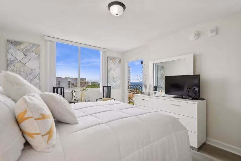 A bright and modern bedroom with a large bed covered in white bedding and pillows. There are two black chairs and decorative wall art near a large window showing a cityscape and blue sky. A white dresser with a mirror and a flat-screen TV is positioned against the wall. A door next to the dresser opens to a balcony with a view of buildings and the ocean in the distance.