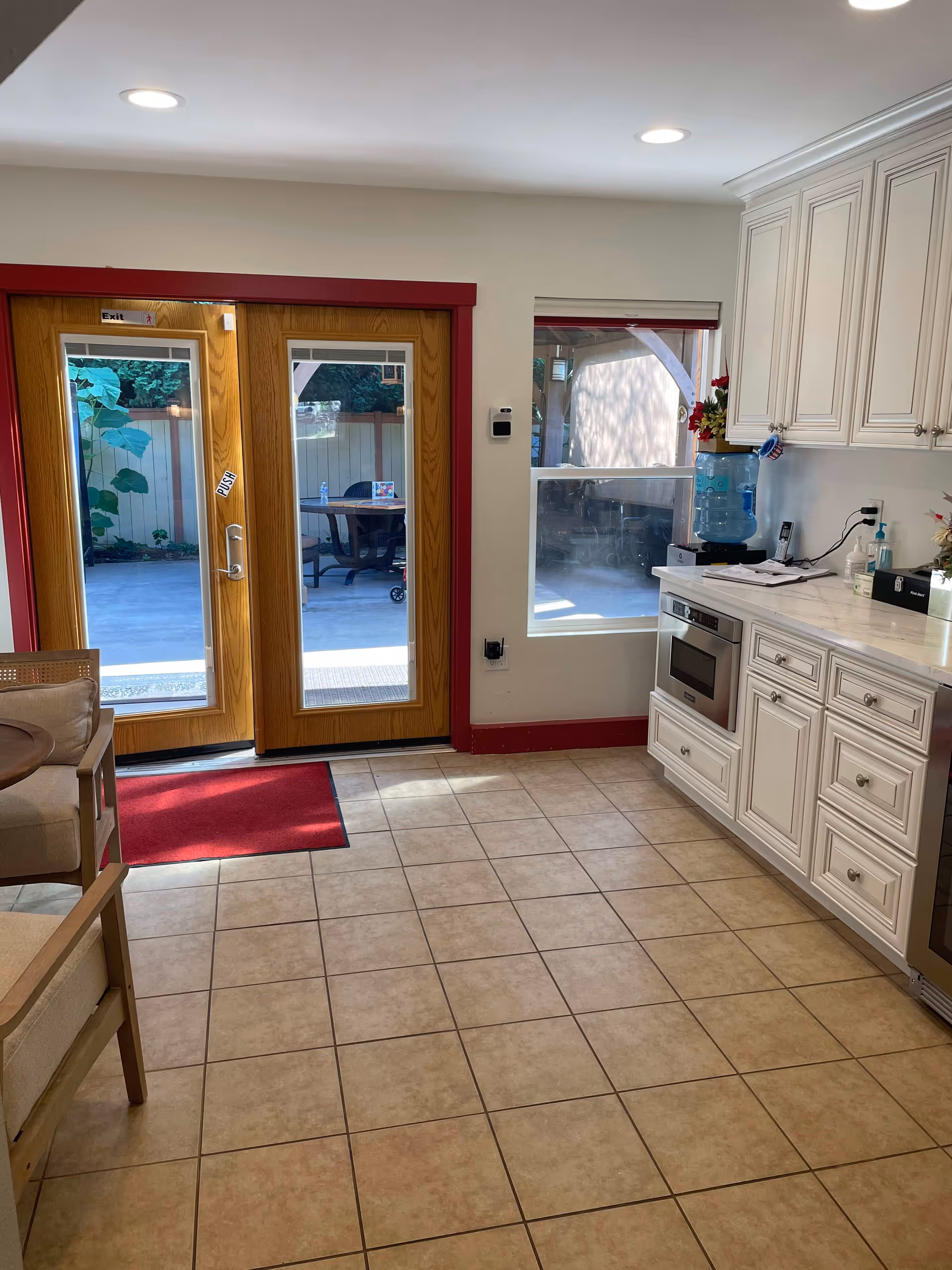 Interior view of a room with beige tiled floor, white cabinetry with a built-in microwave, a water dispenser on the counter, and a window showing an outdoor patio area. There are double wooden doors with glass panels and red trim leading outside, with a red mat in front of the doors. A beige cushioned chair is partially visible on the left side.