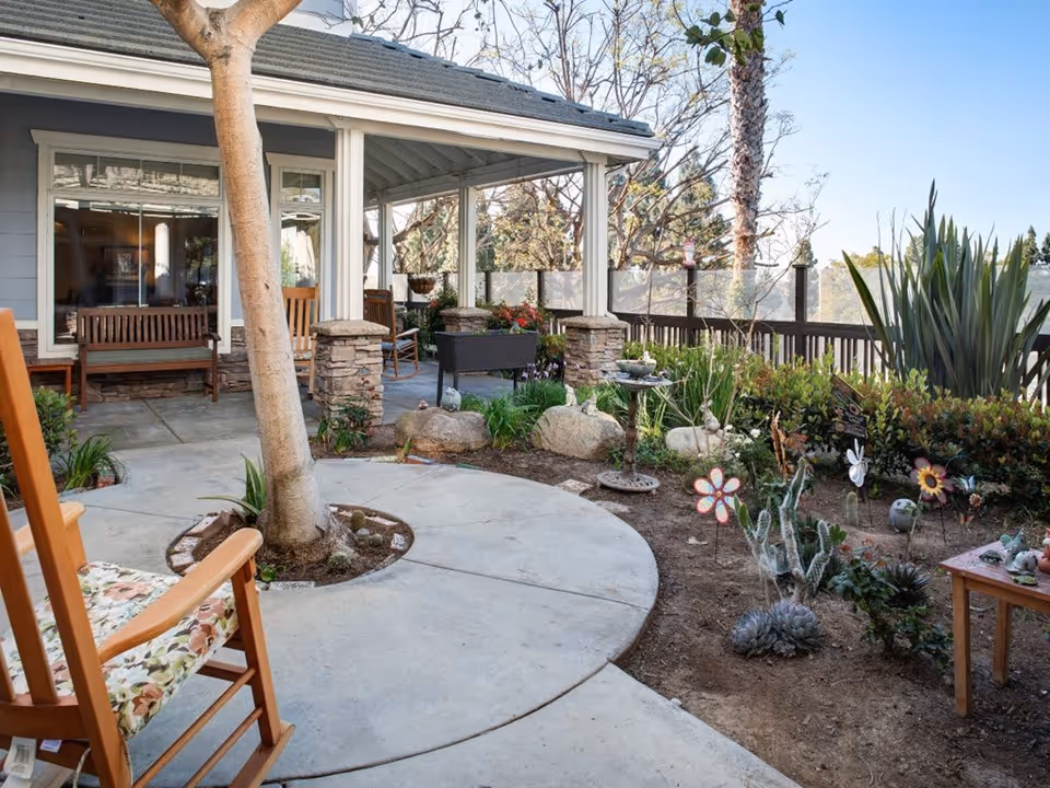 Outdoor patio area at Ivy Park at Huntington Beach featuring a covered seating area with wooden benches and chairs, a circular concrete pathway around a tree, and a garden bed with various plants, flowers, and decorative garden ornaments.