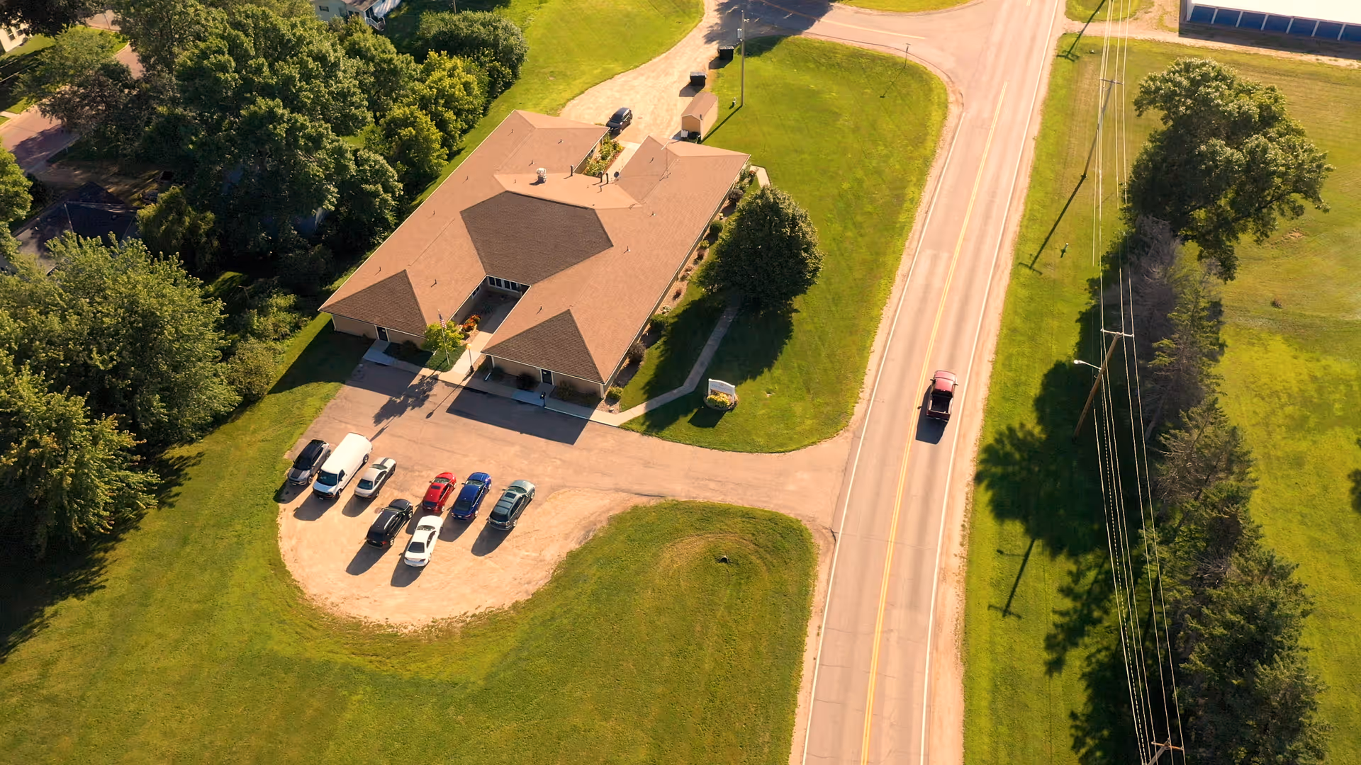 Aerial view of a single-story building with a small parking lot, surrounding lawn, and a road with a passing vehicle.