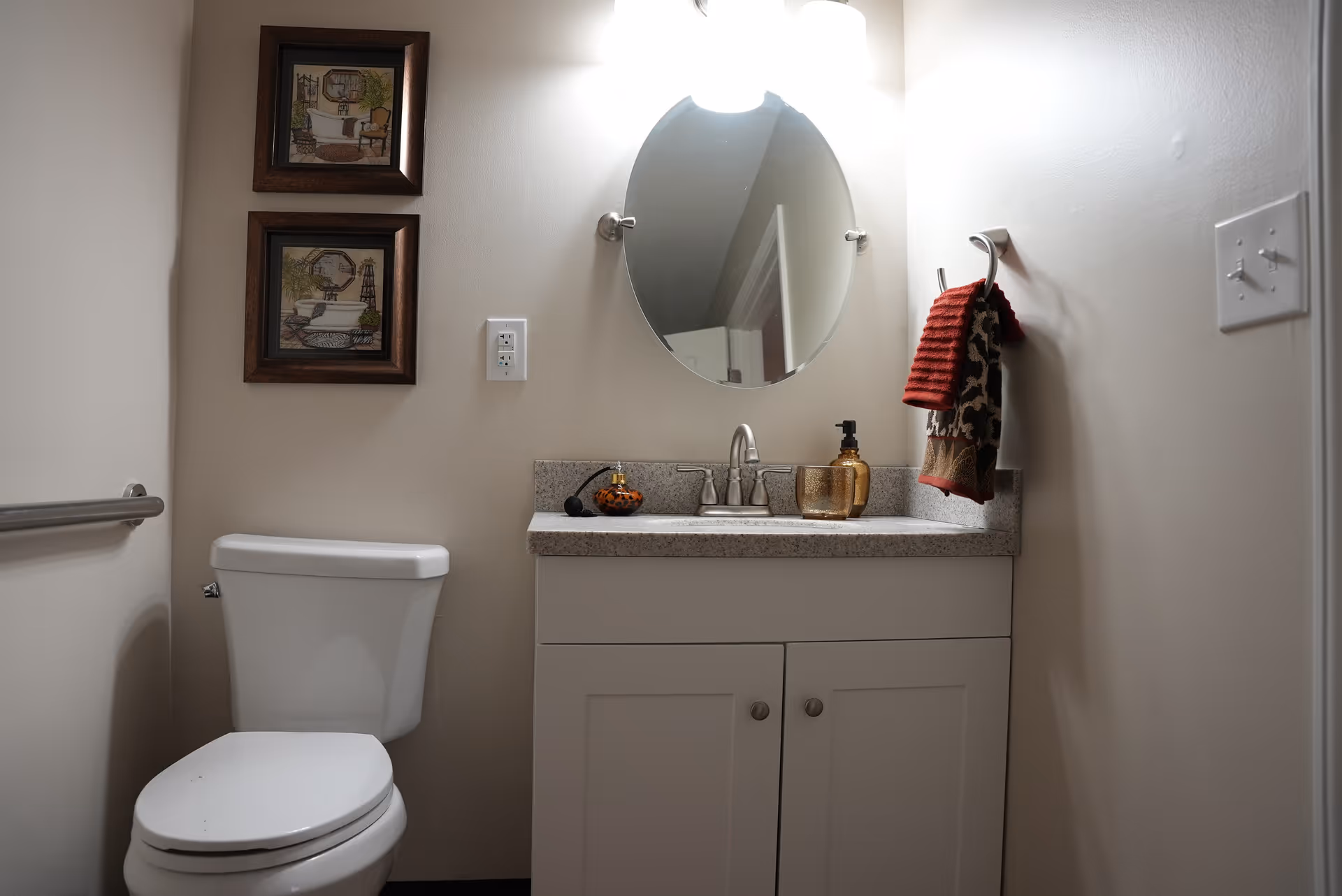 Small bathroom with a toilet, white vanity and countertop sink under an oval mirror, and a towel hanging on the wall.
