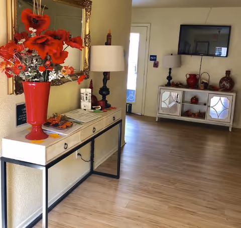 Bright assisted-living lobby with decorative console tables, red floral arrangements, lamps, a mirror, and a wall-mounted TV.