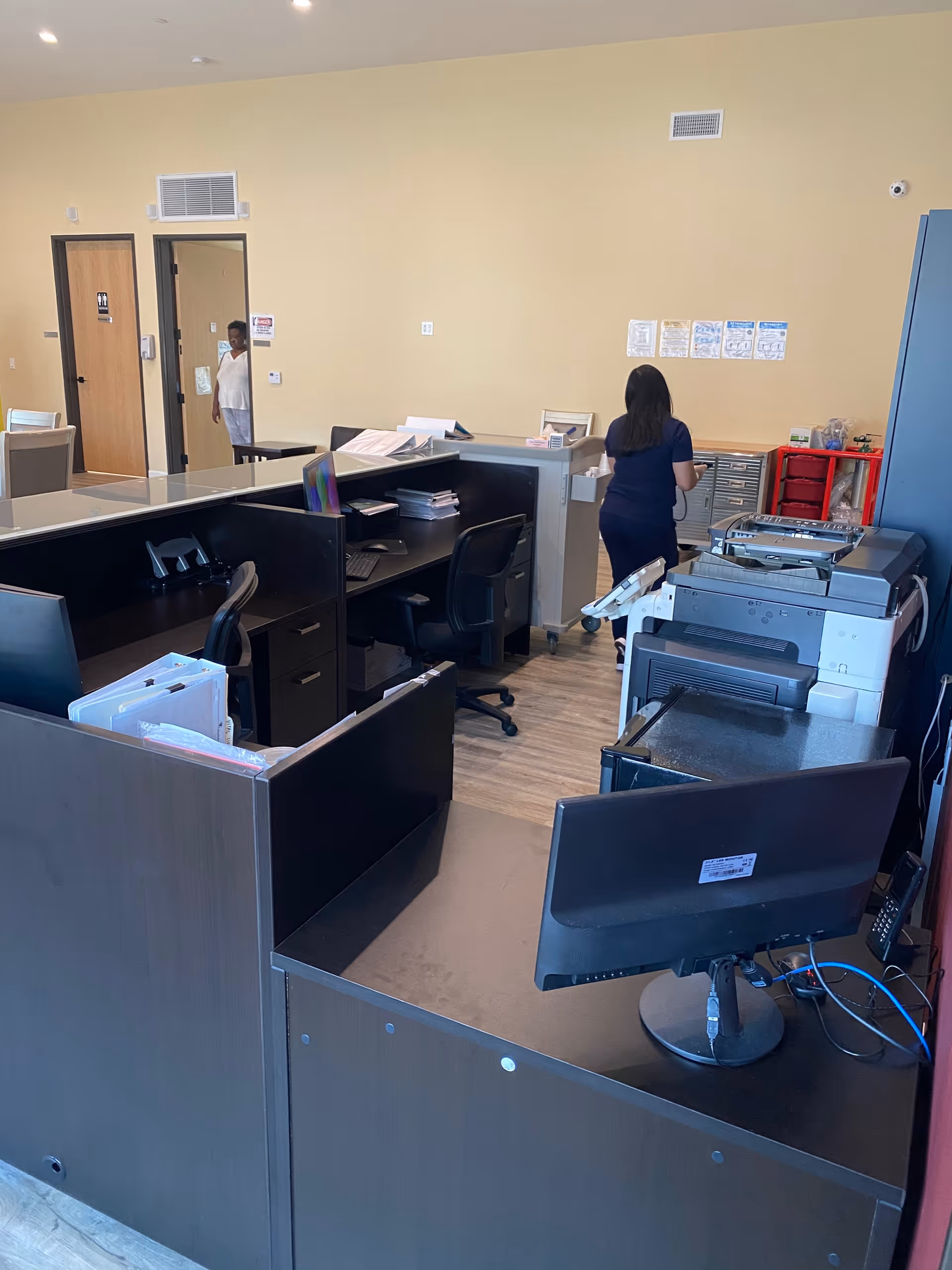 Interior office area with multiple desks, computer monitors, office chairs, and a large printer/copier. A woman is standing near the back wall, and another person is visible through an open door. The walls are beige, and the floor is wood.