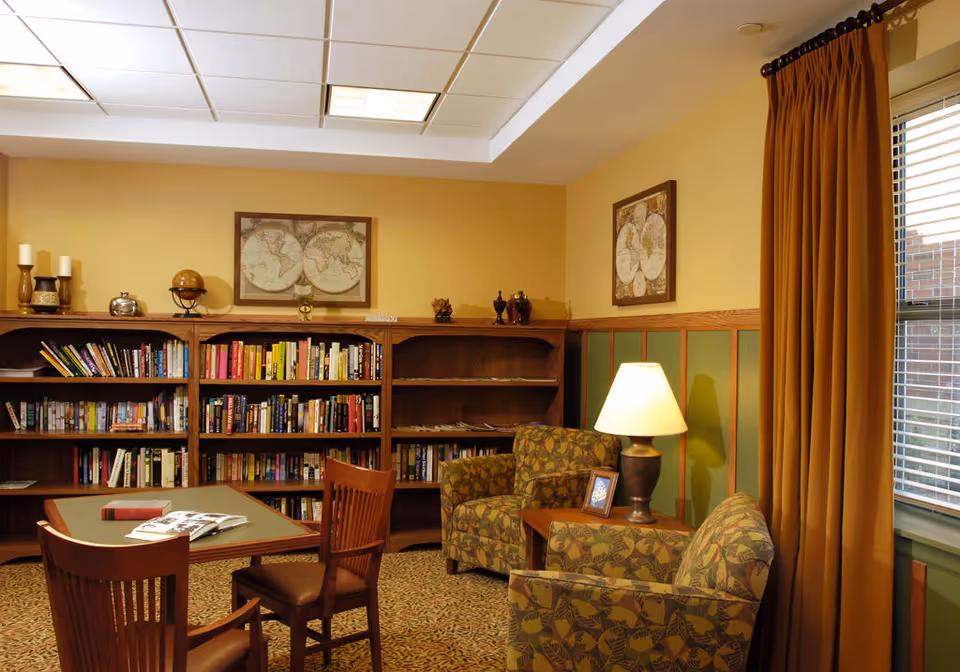 A cozy reading room with wooden bookshelves filled with books, a table with two wooden chairs, two patterned armchairs, a side table with a lamp and a framed photo, yellow walls with green paneling, framed world maps on the walls, and a window with mustard-colored curtains.