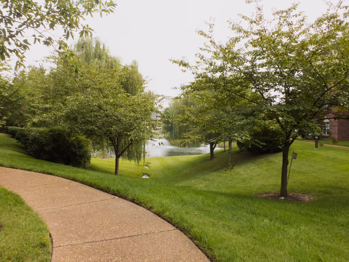 A winding paved pathway surrounded by green grass and trees leading towards a small pond with a fountain in the background, part of a landscaped outdoor area at Blakeford at Green Hills.