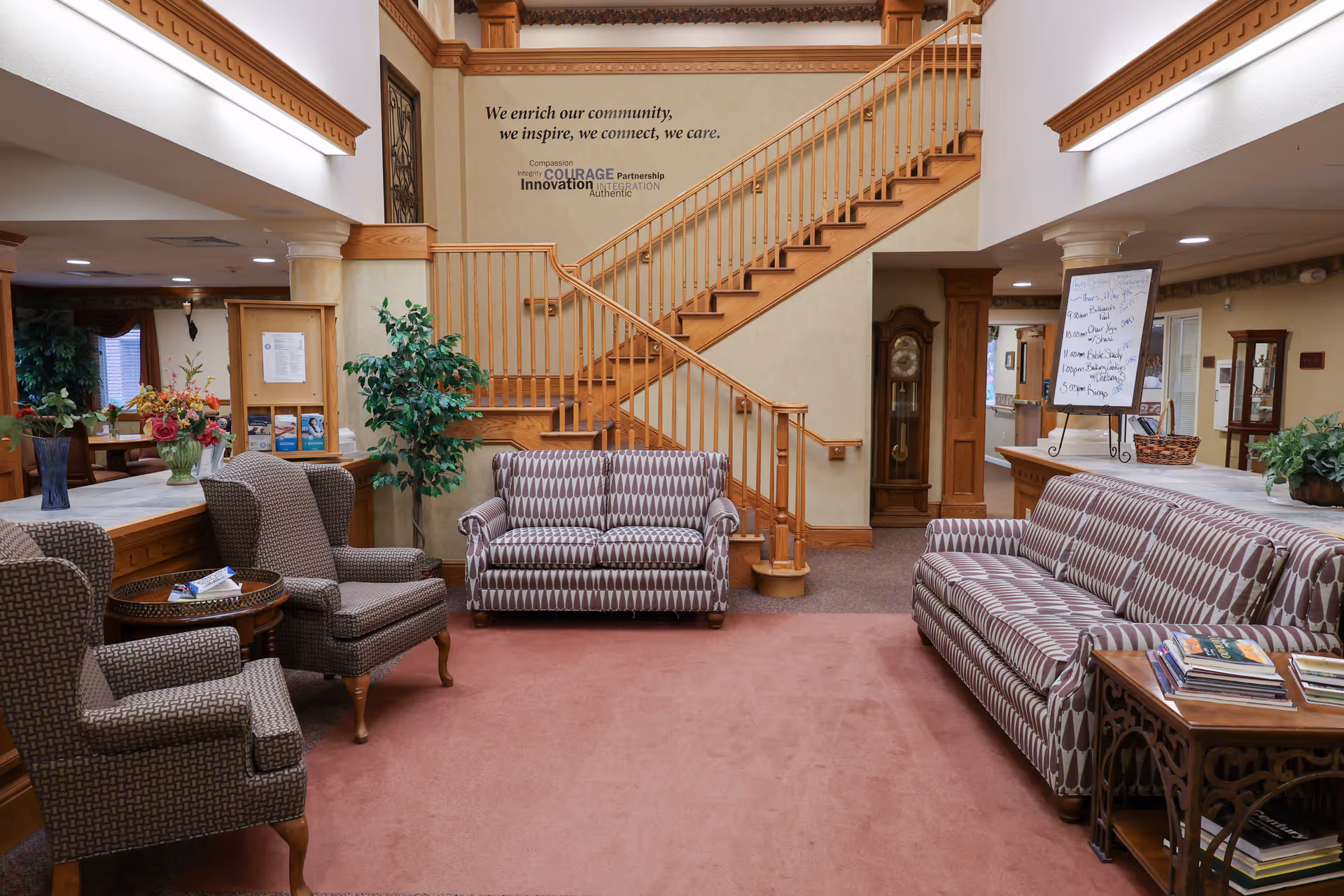 A cozy senior living facility common area with patterned sofas and armchairs arranged around a carpeted floor. A wooden staircase with railings leads to an upper floor. There are plants, a grandfather clock, and a bulletin board with a schedule on an easel. The walls have inspirational text about community and care.