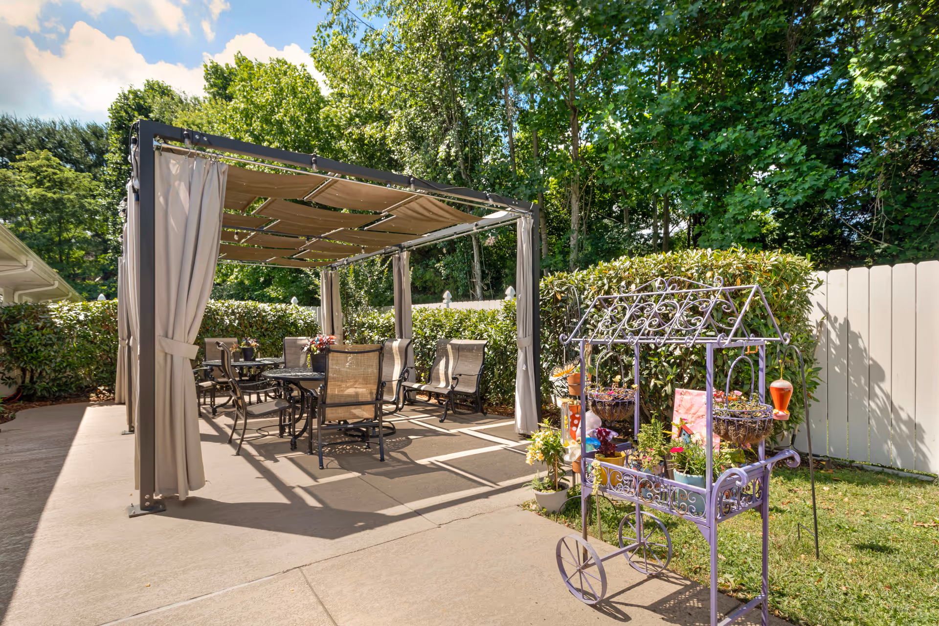 Outdoor patio area with a shaded pergola covering a table and several chairs. There is a decorative purple metal plant stand with various potted plants and flowers on the right side. The area is surrounded by green bushes and trees, with a white fence in the background under a partly cloudy sky.