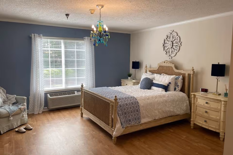 A cozy bedroom with a large bed featuring a woven headboard and footboard, white and blue bedding, two matching nightstands with lamps, a decorative wall piece above the bed, a window with white curtains, a blue accent wall, a comfortable armchair with a blanket, and wooden flooring.
