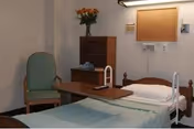 A simple, clean bedroom in a senior living facility featuring a single bed with white sheets and a light blue blanket, a wooden overbed table, a green upholstered chair, a wooden dresser with a vase of orange flowers on top, and a bulletin board on the wall.
