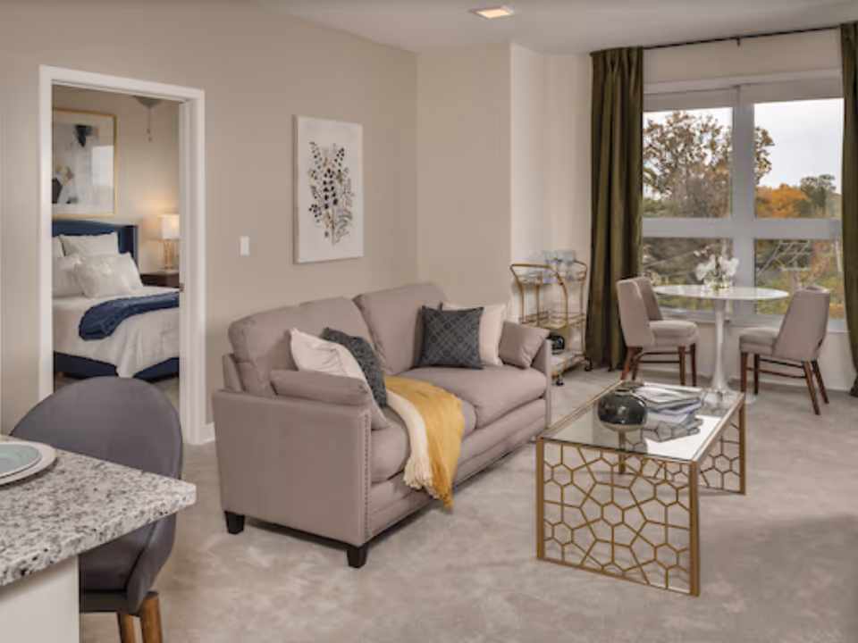 Contemporary living room featuring a beige sofa with throw pillows, a glass-top coffee table, a small dining nook by a large window, and a view into an adjacent bedroom.