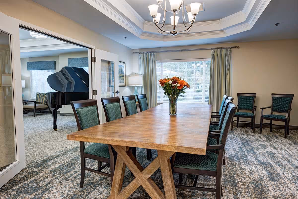 A well-lit dining room with a long wooden table surrounded by green upholstered chairs. A vase with orange and white flowers is placed in the center of the table. In the background, there is a large window with curtains and a chandelier hanging from the ceiling. Through an open door, a black grand piano and additional seating are visible in an adjacent room.