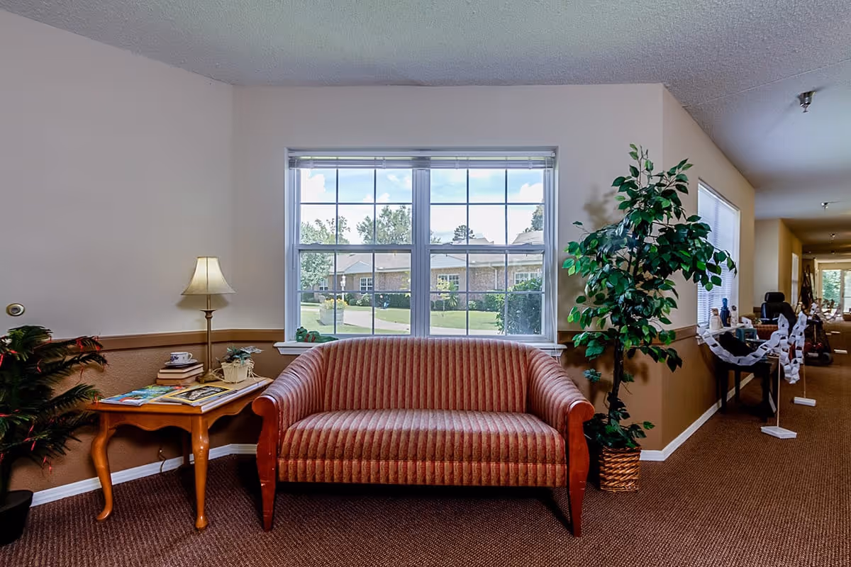 A cozy seating area in a senior living facility featuring a striped upholstered sofa in front of a large window with a view of a garden and buildings outside. To the left of the sofa is a wooden side table with a lamp, books, and a small plant. To the right is a tall potted plant. The area has beige walls with a brown wainscot and carpeted floor, with a hallway extending to the right.