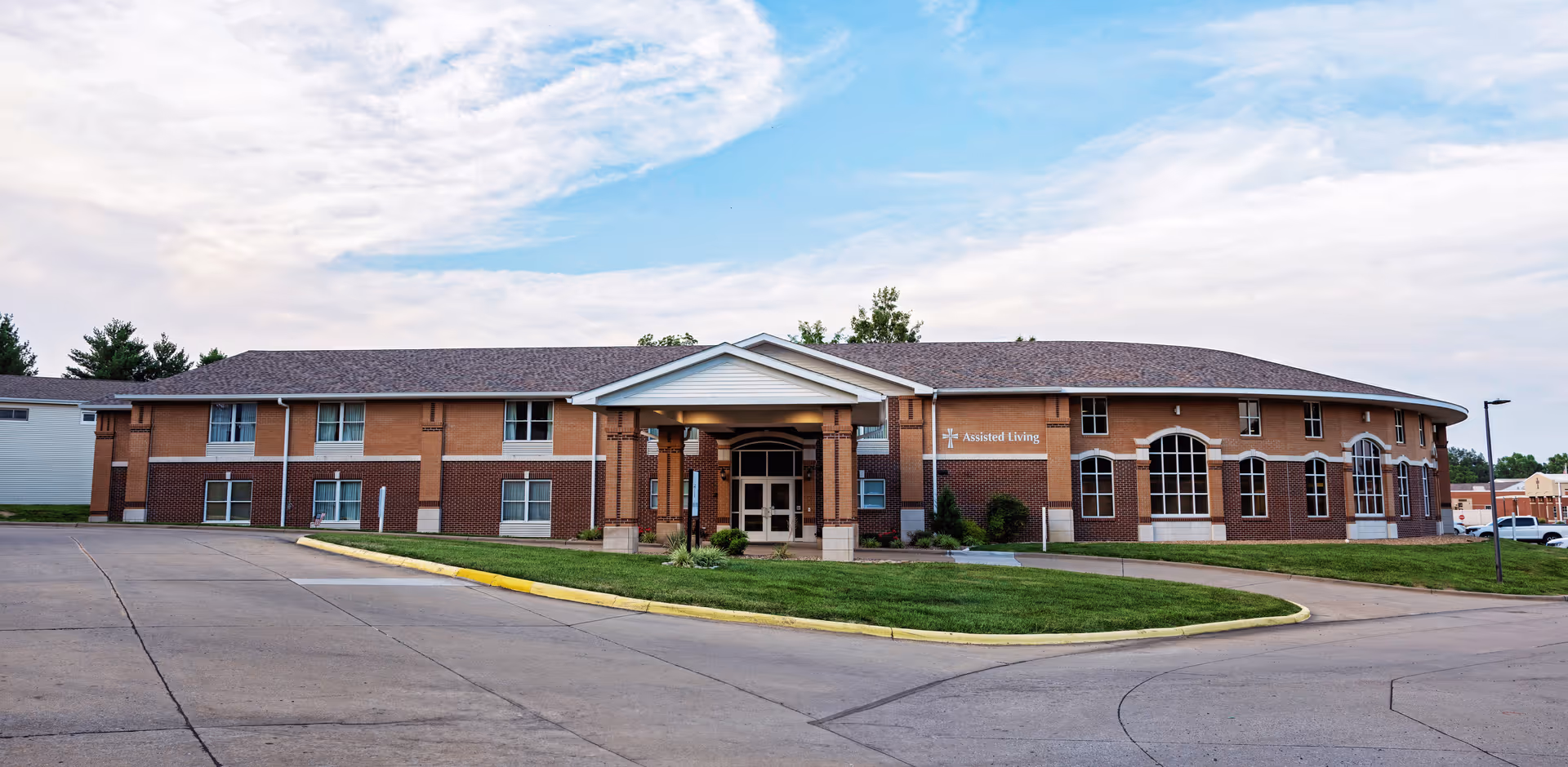 Exterior view of a two-story assisted living facility building with a covered entrance, brick and siding facade, multiple windows, and a curved section on the right side. The building is surrounded by a paved driveway and green lawn under a partly cloudy sky.