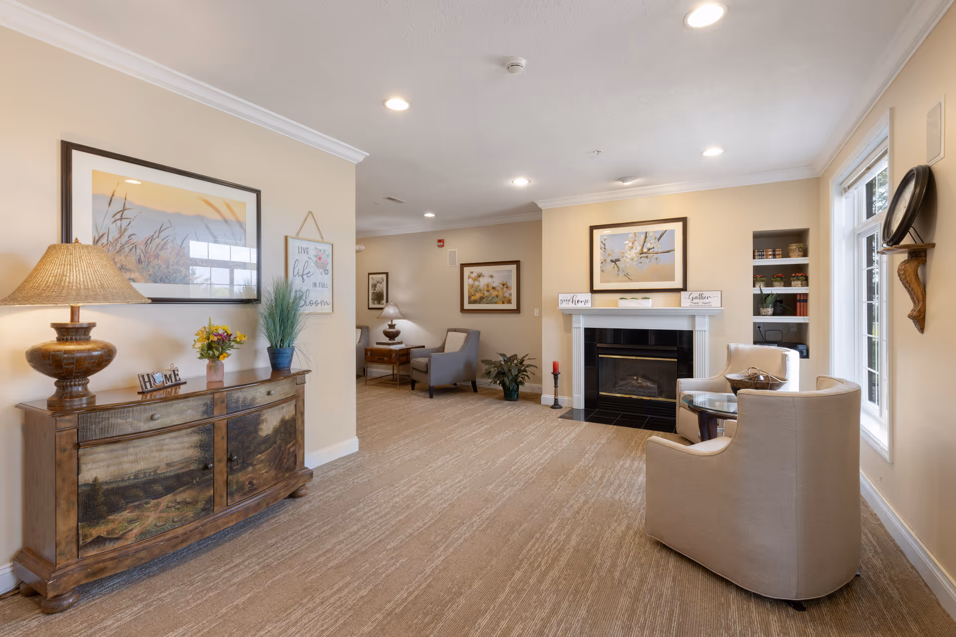 A cozy living room area in a senior living facility with beige walls and carpet. The room features a wooden sideboard with a lamp, flowers, and decorative items, a fireplace with framed artwork above it, two beige armchairs around a glass-top table near a window, and additional seating with armchairs and side tables in the background. The space is warmly lit with recessed ceiling lights.