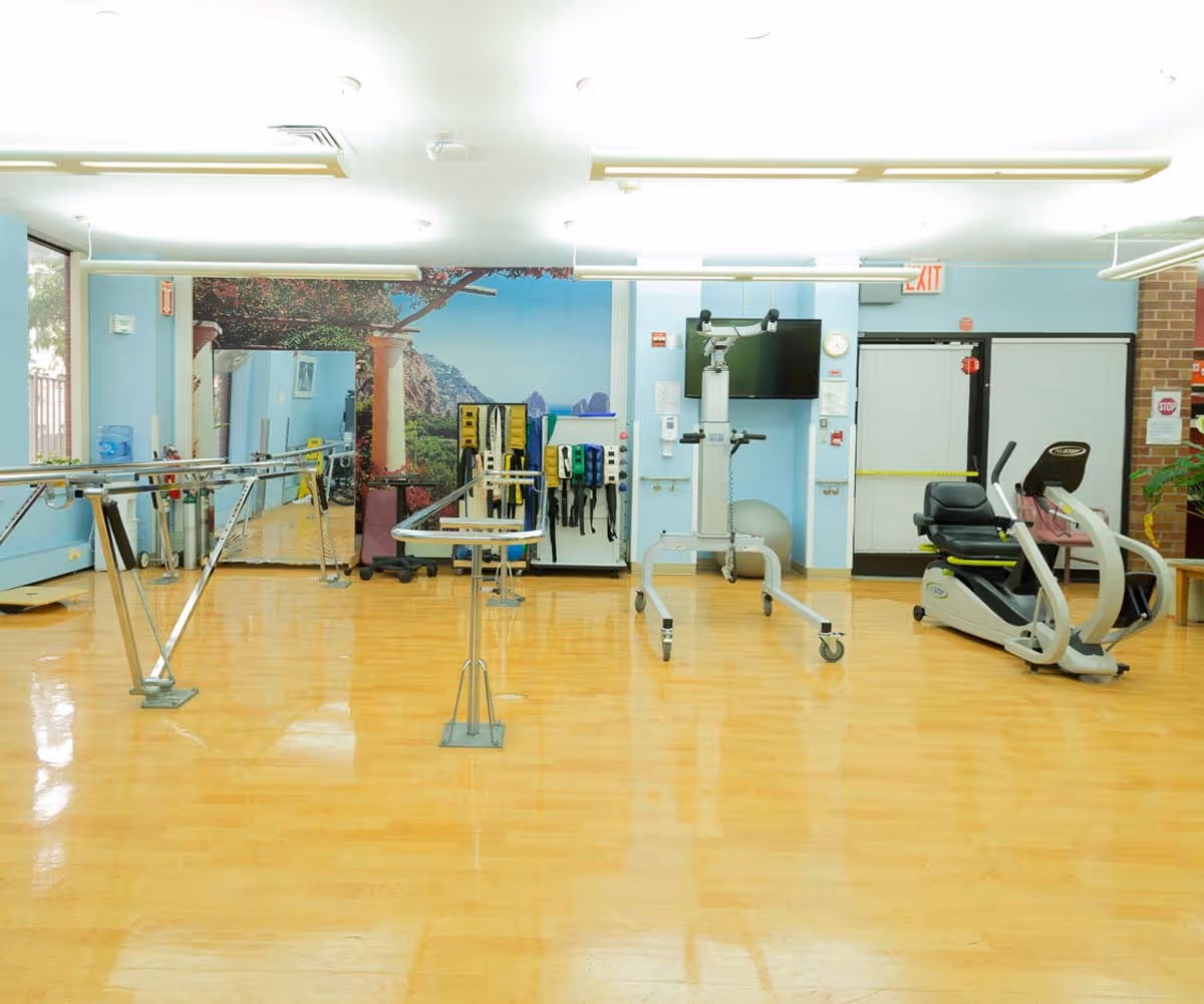 Physical therapy room with parallel bars for walking exercises, exercise equipment including a recumbent bike, a large mirror on the wall, and a mural depicting an outdoor scenic view with columns and greenery.