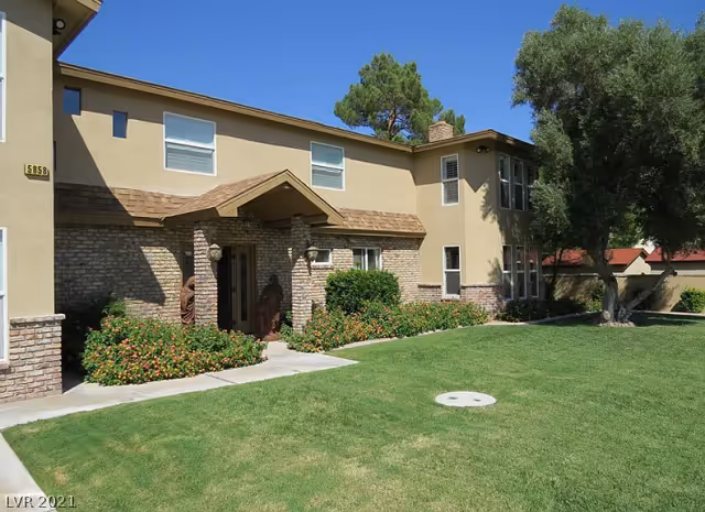 Exterior view of a two-story residential building with beige walls and brick accents. The entrance has a small covered porch supported by brick columns, surrounded by green bushes and flowering plants. There is a well-maintained lawn in the foreground and a large tree on the right side under a clear blue sky.