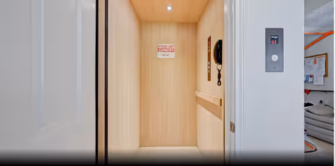 Open elevator with light wood paneling inside, a maximum capacity sign, a telephone on the right wall, and an elevator control panel outside on the right wall showing floor G1. Part of a room with a couch and bulletin board is visible to the right.