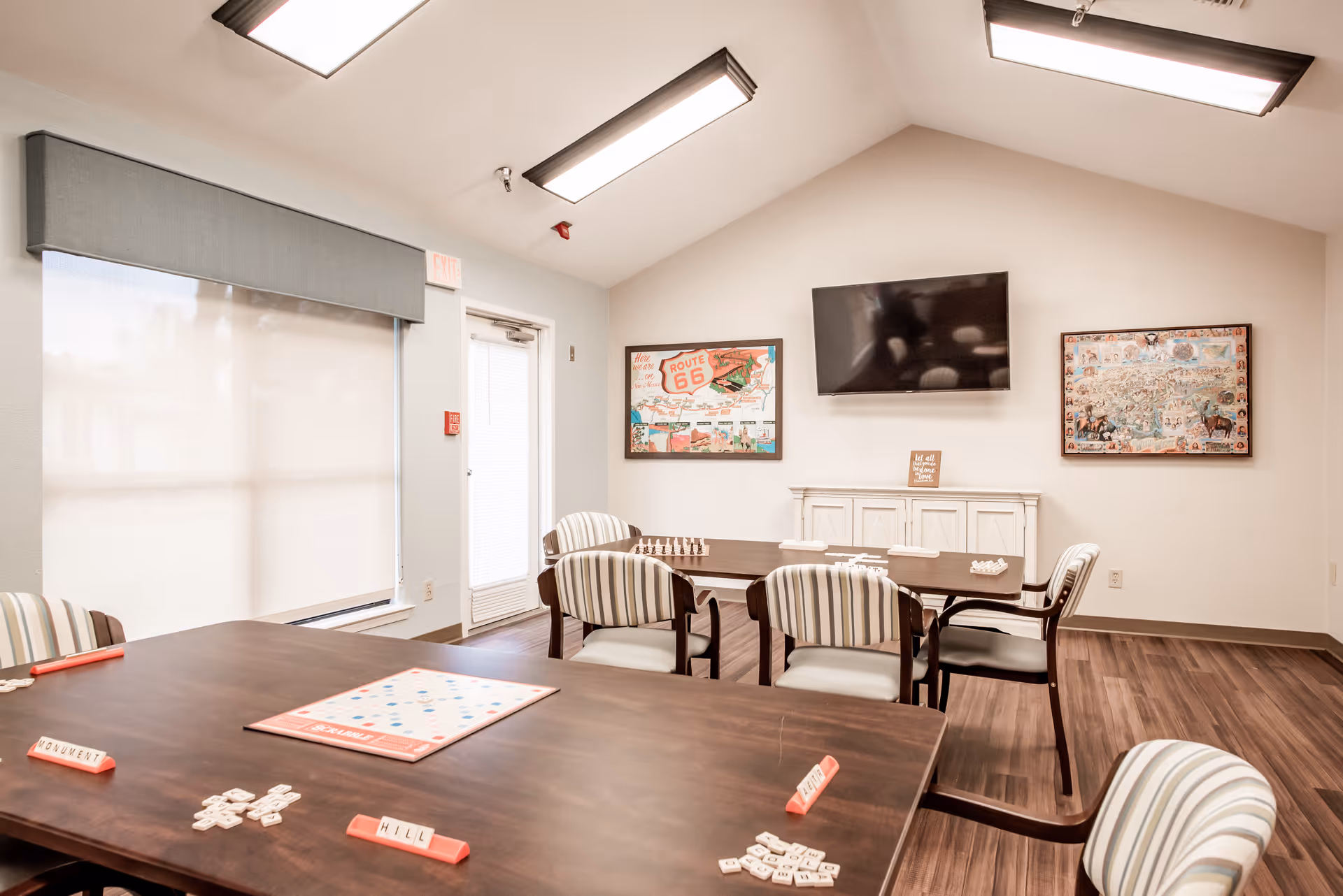 A bright room with several tables and striped cushioned chairs arranged for playing board games. A Scrabble game is set up on the nearest table with letter tiles and racks. The walls are decorated with framed pictures and a flat-screen TV mounted above a white cabinet. Large windows with blinds allow natural light to fill the space.