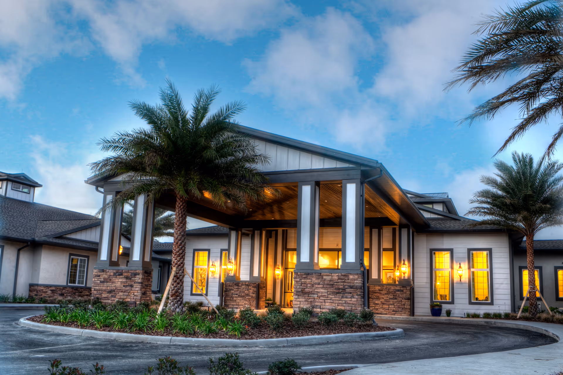 Front entrance of a single-story senior living facility with a covered porte-cochere, lit windows, and palm trees.