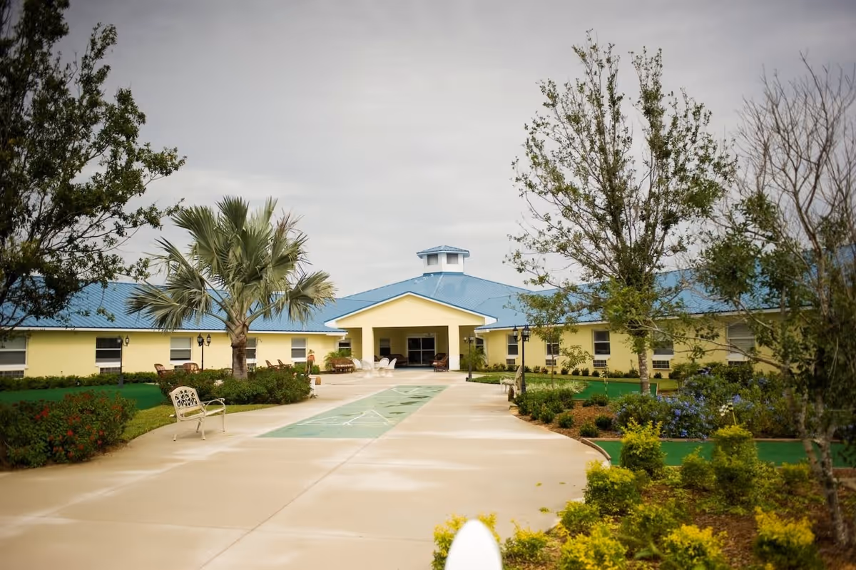 Outdoor view of Southern Life Assisted Living facility with a wide concrete pathway leading to a yellow building with a blue roof. The pathway is flanked by green lawns, trees, shrubs, and benches. The sky is overcast.
