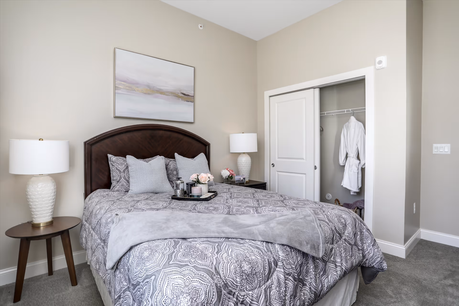 A neatly made bed with patterned gray and white bedding, two gray pillows, and two decorative pillows. There is a tray with flowers, a candle, and a French press on the bed. On either side of the bed are nightstands with white lamps. A closet with sliding doors is partially open, revealing a white robe hanging inside. A framed abstract painting hangs on the beige wall above the bed.