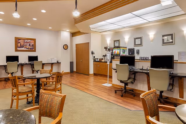 A well-lit common area with several computer workstations along the walls, each equipped with a chair and desktop computer. There are round tables with chairs in the center of the room, and a beverage station with coffee dispensers and cups is visible in the background. The room has wood flooring and carpeted areas, with framed artwork and a clock on the walls.