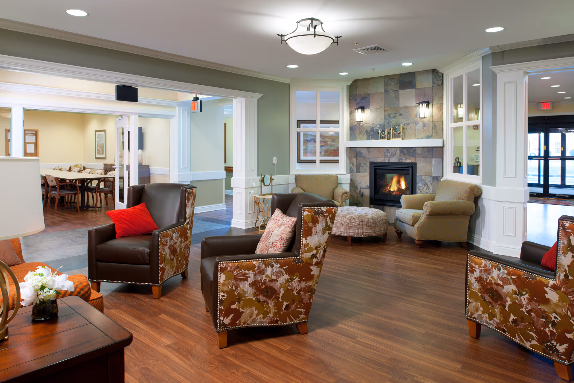 A cozy senior living facility common area with several upholstered armchairs arranged around a fireplace with a tiled surround. The room has wood flooring, soft lighting, and a small ottoman near the fireplace. In the background, there is a dining area with tables and chairs visible through an open doorway.