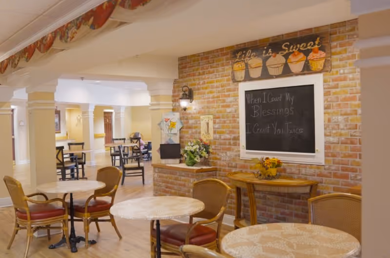 A cozy indoor seating area with round tables and wicker chairs with red cushions. The walls are decorated with a brick accent wall featuring a chalkboard with the message 'When I Count My Blessings I Count You Twice' and a sign above it that says 'Life is Sweet' with cupcake illustrations. The space has warm lighting and an inviting atmosphere with additional tables and chairs visible in the background.