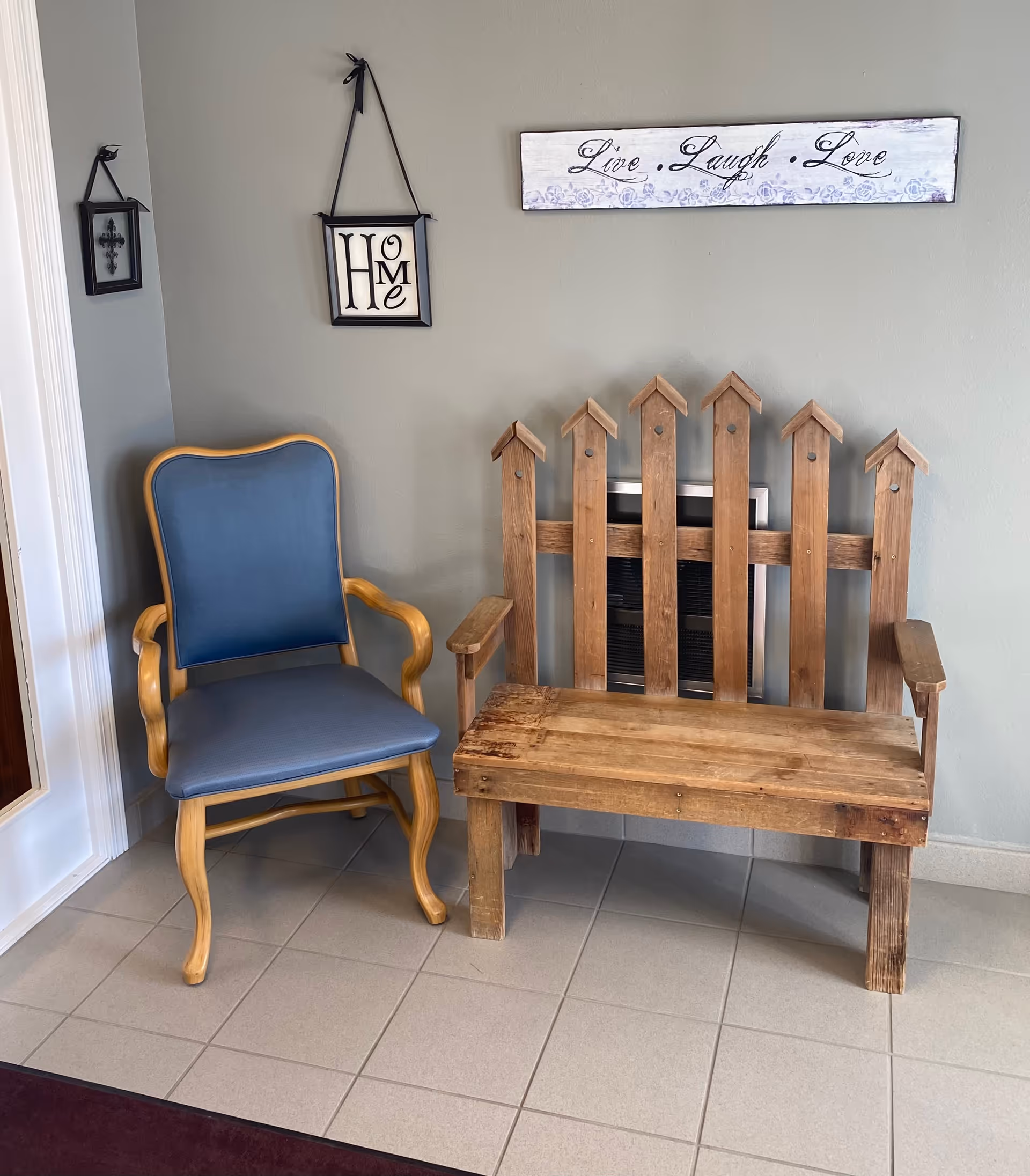 A small seating area with a blue cushioned wooden armchair and a rustic wooden bench designed like a picket fence backrest, placed against a light gray wall. Above the bench is a decorative sign that reads 'Live Laugh Love' and to the left is a hanging framed decoration with the word 'Home'. The floor is tiled in light gray.