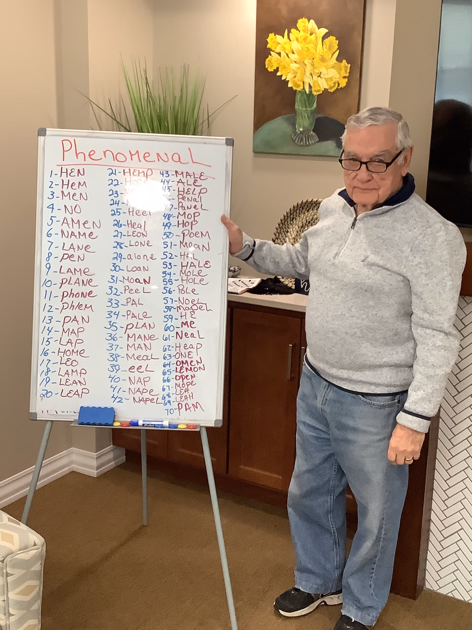An elderly man wearing glasses, a light gray pullover, and blue jeans stands next to a whiteboard on an easel. The whiteboard has the word 'Phenomenal' written at the top in red marker, with a long list of words written underneath in blue and red marker. Behind him is a cabinet with a decorative basket and a painting of yellow flowers on the wall.