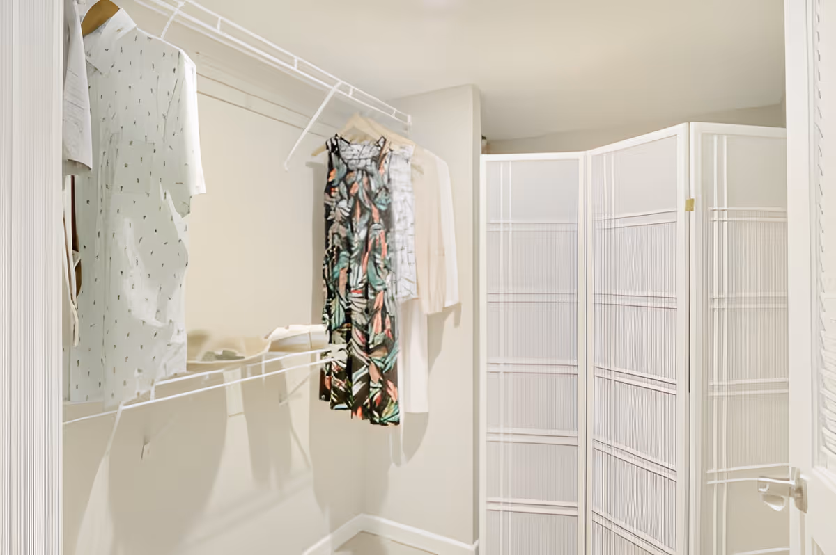 A small walk-in closet with a white wire shelf holding several hanging clothes including a patterned dress and light-colored shirts. A white folding room divider is positioned in the corner near a white door with a silver handle.