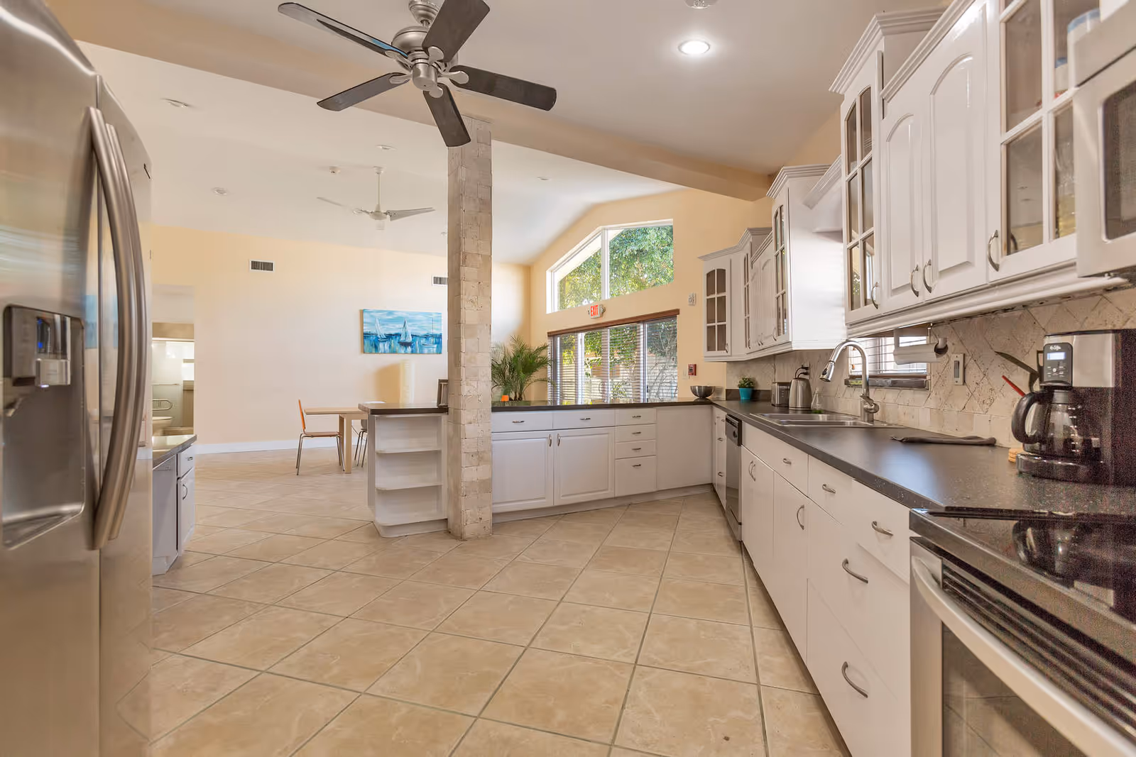 Bright and spacious kitchen area with white cabinets, black countertops, a stainless steel refrigerator, stove, and coffee maker. The kitchen opens into a dining area with a table and chairs, beige tiled floor, ceiling fans, and large windows letting in natural light.